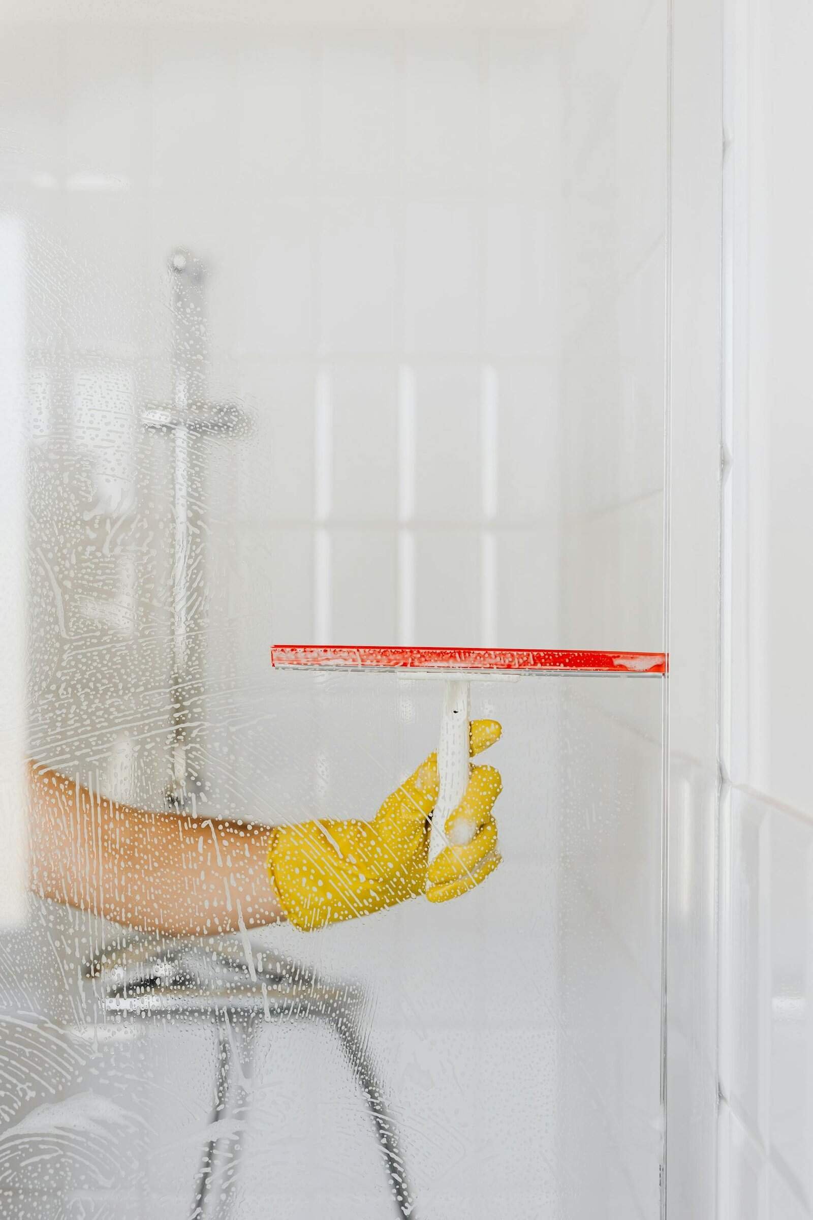 A person wearing a yellow glove cleans a shower glass with a squeegee, emphasizing hygiene and cleanliness.